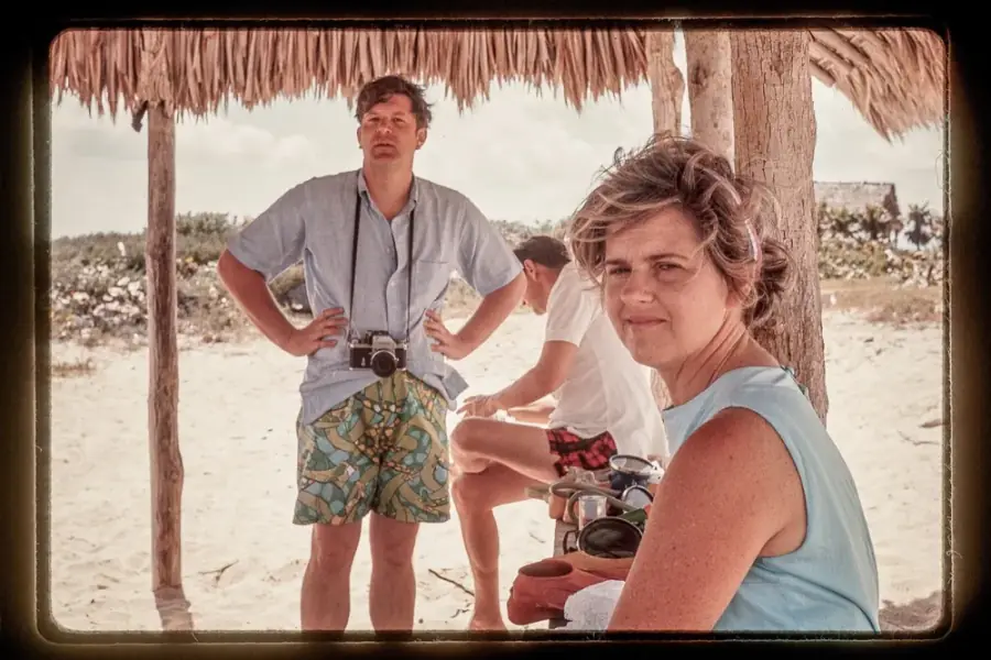 A vintage beach photo: a man with a camera stands under a thatched shelter, a woman sits in the foreground; Standard AI has framed both faces
